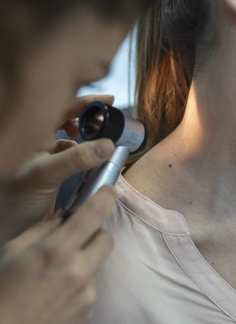 Dermatologist in latex gloves holding dermatoscope while examining attractive patient with skin disease. Female dermatologist examining patient with dermascope, looking for signs of skin cancer.
